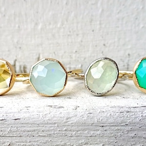 Op de afbeelding: Vier ringen met verschillende gekleurde edelstenen. De ringen zijn van goud en zilver met gefacetteerde edelstenen. De edelstenen zijn geel, lichtgroen, wit en groen.