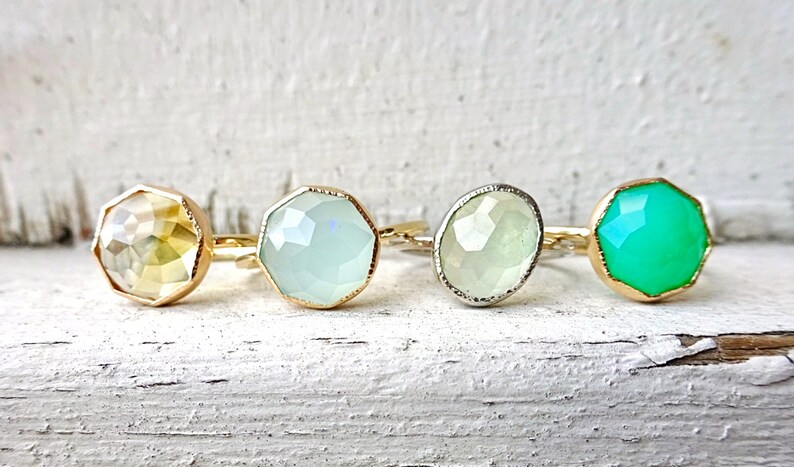 Op de afbeelding: Vier ringen met verschillende gekleurde edelstenen. De ringen zijn van goud en zilver met gefacetteerde edelstenen. De edelstenen zijn geel, lichtgroen, wit en groen.