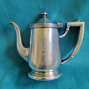 May include: A silver-colored, vintage coffee pot with a curved spout and a handle. The pot has a lid and a decorative emblem on the front. The pot is sitting on a teal surface.