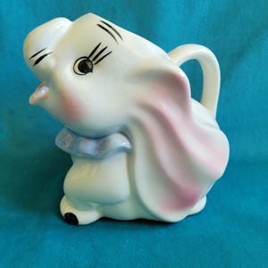 May include: A white ceramic elephant creamer with a handle and a blue collar. The elephant has pink inner ears, black eyelashes, and a smiling expression. The creamer is a whimsical, decorative piece.