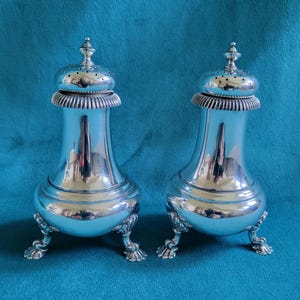 May include: Two silver salt and pepper shakers with a decorative design and three legs each. The shakers have a shiny finish and are on a blue background.