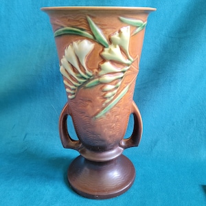 May include: A brown and green glazed ceramic vase with two handles and a floral design. The vase features a stylized depiction of white flowers with green leaves.