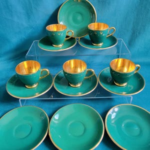 May include: A set of six green and gold teacups and saucers. The cups and saucers have a gold rim and a green interior. The cups are all the same size and shape. The saucers are all the same size and shape.