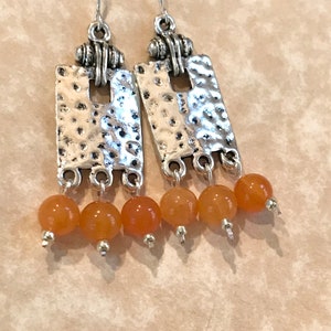 May include: A pair of silver-toned dangle earrings. Each earring features a textured rectangular plate with a small square cutout, and three orange-colored beads. The earrings have a hook closure.