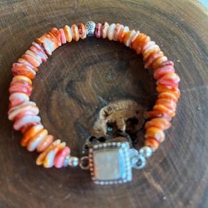 Könnte beinhalten: Ein Armband aus orangefarbenen und weißen Muschelkernperlen. Es hat einen quadratischen, silberfarbenen Verschluss mit einem weißen Stein. Die Perlen sind mit silberfarbenem Metall aufgefädelt. Das Armband liegt auf einer Holzoberfläche.