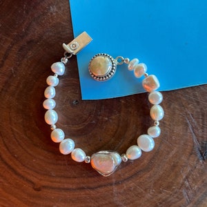 May include: A white pearl bracelet with a heart-shaped pearl and a round pearl charm. The bracelet is on a wooden surface.