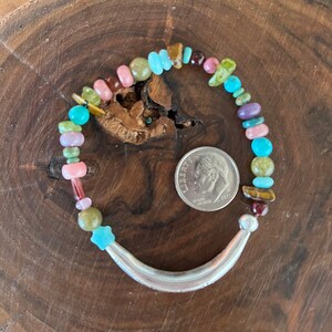 May include: A colorful beaded bracelet with a silver crescent moon charm. The beads are a mix of turquoise, pink, green, and brown.