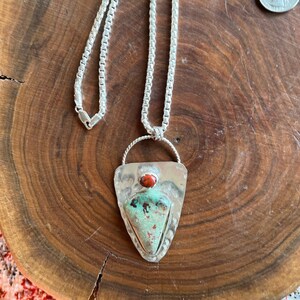 May include: A silver chain necklace with a triangular pendant featuring a turquoise stone and a red stone accent. The pendant is set in a silver frame with a textured design.