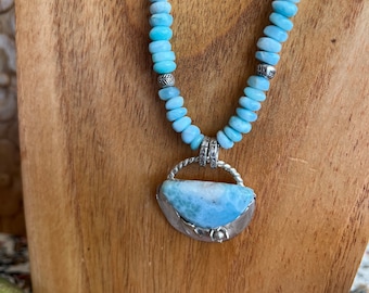May include: A light blue gemstone necklace with a silver pendant. The pendant features a large, crescent-shaped gemstone with a silver setting. The necklace is made with small, round beads and a silver chain.
