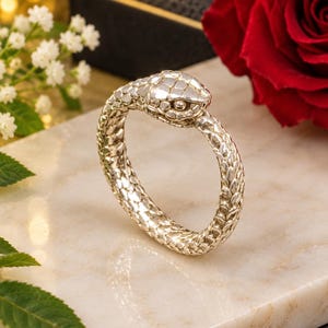 May include: A silver-toned bracelet shaped like a coiled snake with detailed scale textures covering the entire surface. The snake head is positioned at the top, featuring defined eyes and a smooth, polished finish that reflects light across the metal.