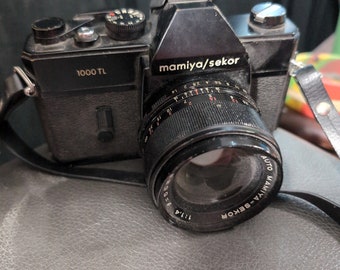 May include: A black Mamiya Sekor 1000 TL film camera with a silver lens. The lens has a black ring with white markings and the text "AUTO MAMIYA-SEKOR 1:1.4 f=55mm" printed on it.