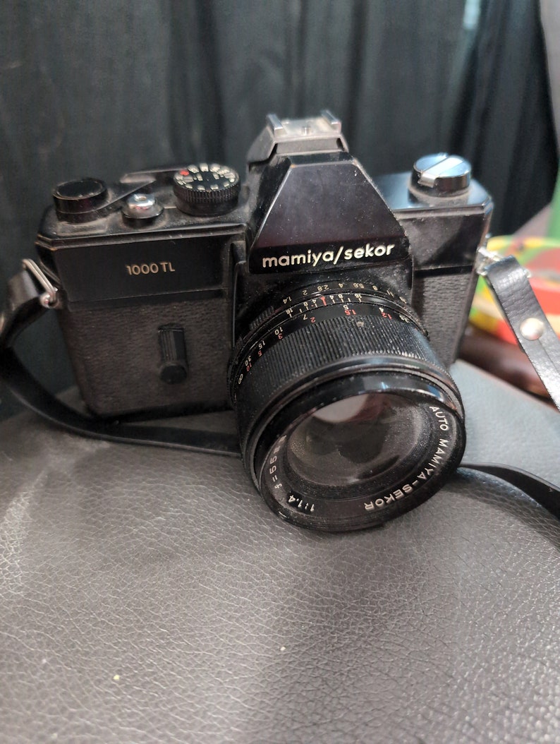 K&ouml;nnte beinhalten: Eine schwarze Mamiya Sekor 1000 TL Filmkamera mit einem silbernen Objektiv. Das Objektiv hat einen schwarzen Ring mit wei&szlig;en Markierungen und den Text "AUTO MAMIYA-SEKOR 1:1.4 f=55mm" aufgedruckt.