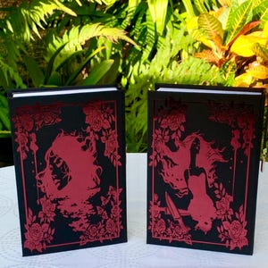 May include: Two black hardcover journals with red floral designs and a silhouette of a woman on the front cover.