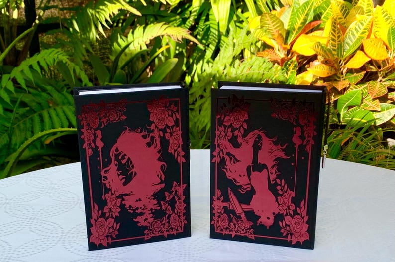 May include: Two black hardcover journals with red floral designs and a silhouette of a woman on the front cover.