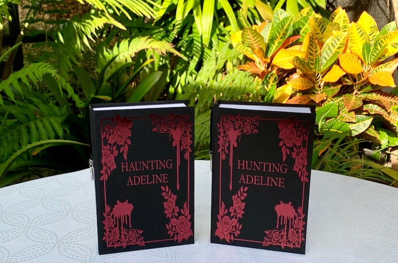 May include: Two black hardcover books with red floral designs and the title "Haunting Adeline" and "Hunting Adeline" on the front covers.
