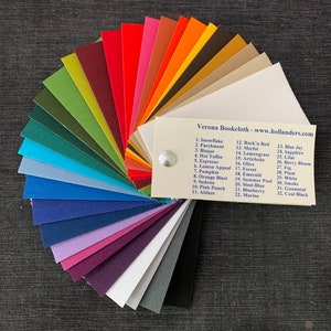 May include: A color chart of 32 fabric swatches in a fan shape. The swatches are labeled with numbers and names, such as Snowflake, Rock'n Red, and Blue Jay. Verona Bookcloth - www.hollanders.com is printed on the chart.