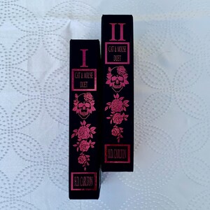 May include: Two black hardcover books with pink and silver foil lettering. The books are titled "Cat & Mouse Duet" and are numbered I and II. The covers feature a skull and roses.