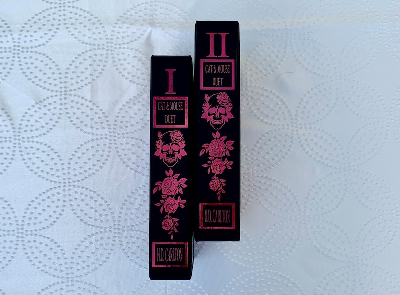 May include: Two black hardcover books with pink and silver foil lettering. The books are titled "Cat & Mouse Duet" and are numbered I and II. The covers feature a skull and roses.