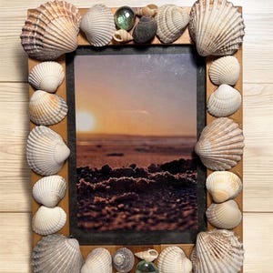 May include: Wooden picture frame adorned with assorted seashells. The frame holds a photo of a sunset over the sea. The shells are in shades of white, beige, and brown, creating a seaside aesthetic. The frame measures approximately 20 cm x 15 cm.