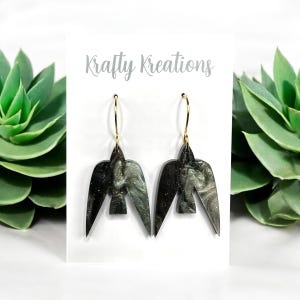 May include: Bird-shaped earrings with gold hoops. The earrings are black with a marbled grey and silver design. The text "Krafty Kreations" is above the earrings.