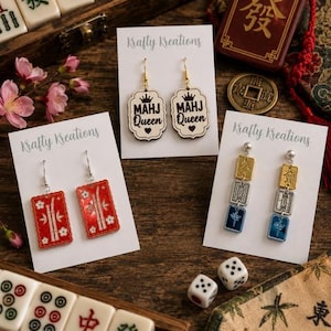 Könnte beinhalten: Eine Auswahl an Ohrringen mit Mahjong-Motiv, präsentiert auf weißen Karten mit dem Logo "Krafty Kreations". Die Ohrringe zeigen rote rechteckige Designs, "MAHJ Queen" Designs und goldene, silberne und blaue Elemente. Mahjong-Steine und Würfel sind ebenfalls im Bild zu sehen.