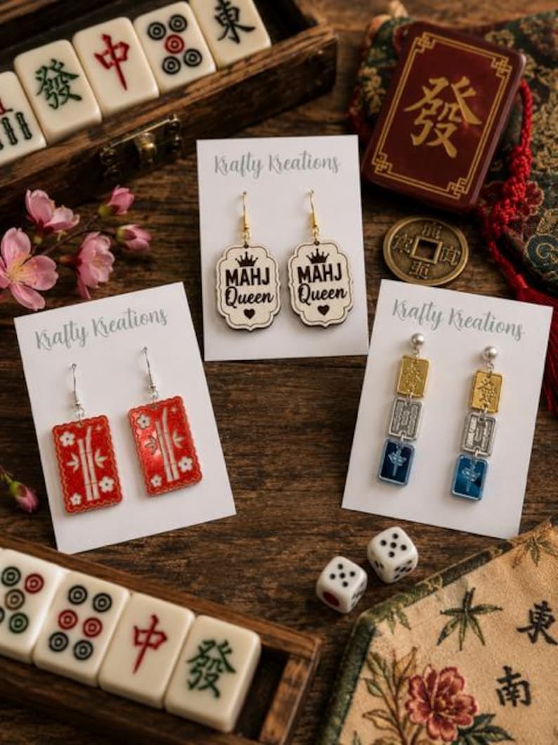 May include: A collection of earrings with a Mahjong theme. The earrings are displayed on white cards with the "Krafty Kreations" logo. The earrings include red rectangular designs, "MAHJ Queen" designs, and gold, silver, and blue designs. Mahjong tiles and dice are also visible.