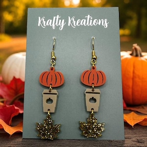 May include: A pair of dangle earrings with a fall theme. Each earring features a gold-colored hook, an orange pumpkin, a tan coffee cup, and a gold glittery maple leaf. The earrings are displayed on a gray card with the text "Krafty Kreations".