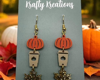 May include: A pair of dangle earrings with a fall theme. Each earring features a gold-colored hook, an orange pumpkin, a tan coffee cup, and a gold glittery maple leaf. The earrings are displayed on a gray card with the text "Krafty Kreations".