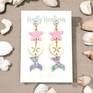 Puede incluir: Un par de pendientes colgantes con temática de sirena. Cada pendiente presenta una estrella de mar rosa, un dólar de arena color crema y una cola de sirena brillante. Los pendientes están suspendidos de una perla y herrajes dorados. El texto "Kratty Kreations" es visible.