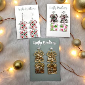 May include: Three pairs of festive earrings are displayed. The earrings are in the shape of gift boxes and bows. The earrings are in red, green, and white, gold, and pink. The brand name "Krafty Kreations" is printed on the cards.
