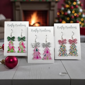 May include: Three pairs of Christmas tree earrings with glittery bows. The earrings are on white cards with the text "Krafty Kreations". The tree designs vary in color and pattern, including pink, green, and confetti.