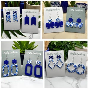 May include: A selection of handmade earrings in blue and white. The earrings showcase floral patterns and solid blue acrylic in diverse shapes, including bows, rectangles, and circles. Each pair is presented on a white or grey card, ideal for gifting.