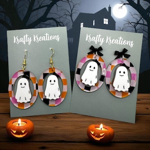 May include: Halloween-themed earrings featuring a white ghost design within an oval frame with a checkered pattern in black, orange, and pink. The earrings are displayed on gray cards with the text "Krafty Kreations". Two lit jack-o'-lanterns are in the foreground.