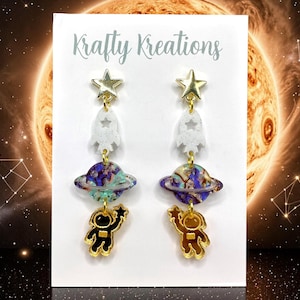 Può includere: Un paio di orecchini pendenti su un cartoncino bianco con la scritta Krafty Kreations. Ogni orecchino presenta una stella dorata, un razzo bianco maculato, un pianeta marmorizzato viola e blu con anello e un astronauta color oro.