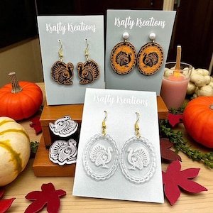 May include: Three pairs of earrings with turkey designs are displayed. One pair is wooden with a turkey cutout, another has a turkey design on an oval with a pearl, and the third is clear with a turkey design. The text "Krafty Kreations" is visible.