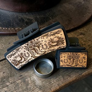 May include: Two black hair clips with a wooden design featuring a cowboy boot, horseshoe, and floral elements. A small, silver, round container sits in front of the clips. The clips are on a wooden surface.