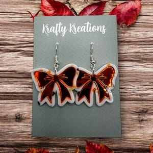 May include: A pair of bow-shaped earrings with a white and tortoiseshell pattern, outlined in white, and a reflective copper-colored center. The earrings hang from silver hooks. The text "Krafty Kreations" is visible on the gray card.