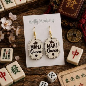 May include: Wooden earrings with the text "MAHJ Queen" and a crown design. The earrings are displayed on a white card with the "Krafty Kreations" logo, surrounded by Mahjong tiles, dice, and a vintage coin on a wooden surface.