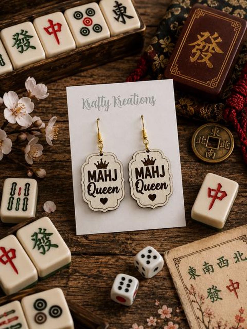 May include: Wooden earrings with the text "MAHJ Queen" and a crown design. The earrings are displayed on a white card with the "Krafty Kreations" logo, surrounded by Mahjong tiles, dice, and a vintage coin on a wooden surface.