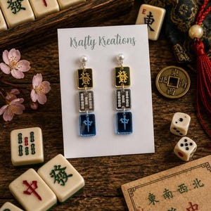 May include: Earrings with a pearl stud, gold and black top, silver middle, and blue bottom. The earrings are displayed on a white card with the text "Krafty Kreations". The image includes Mahjong tiles, dice, and a coin.