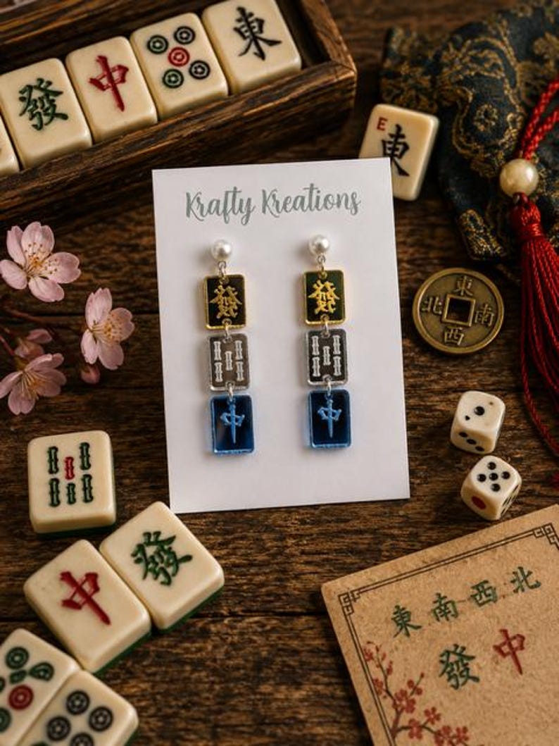 May include: Earrings with a pearl stud, gold and black top, silver middle, and blue bottom. The earrings are displayed on a white card with the text "Krafty Kreations". The image includes Mahjong tiles, dice, and a coin.