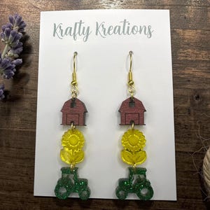 May include: A pair of dangle earrings on a white Krafty Kreations display card. Each earring features a stacked design with a dark red wooden barn at the top, a yellow acrylic sunflower in the middle, and a green glittery tractor at the bottom.