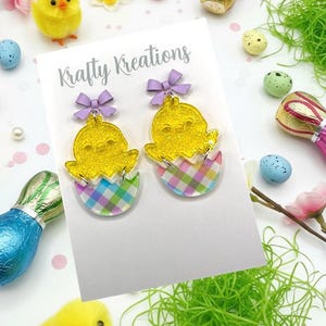 May include: A pair of yellow chick earrings with purple bows, emerging from pastel plaid eggshells. The earrings are on a white card with the text "Krafty Kreations". Easter-themed decorations, including chicks and eggs, surround the earrings.