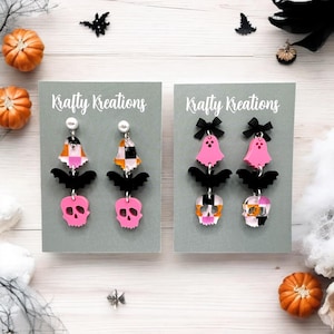 Op de afbeelding: Twee paar Halloween-oorbellen op grijze kaarten. Eén paar heeft roze schedels, zwarte vleermuizen en geblokte belvormen. Het andere paar heeft roze spookvormen, zwarte vleermuizen en geblokte schedelvormen. De kaarten lezen "Krafty Kreations".