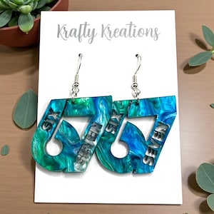 May include: Dangle earrings with a marbled blue and green pattern. Each earring is in the shape of the number 67, with the words "SIX" and "SEVEN" cut out. They hang from silver-coloured hooks. The background includes a white card with the text "Krafty Kreations".