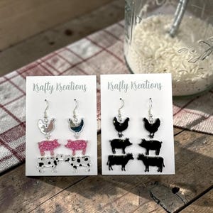 May include: Two pairs of earrings with farm animal designs. One pair features a silver chicken, pink glitter pig, and black and white cow. The other pair has a black chicken, pig, and cow. The text "Krafty Kreations" is on the cards.