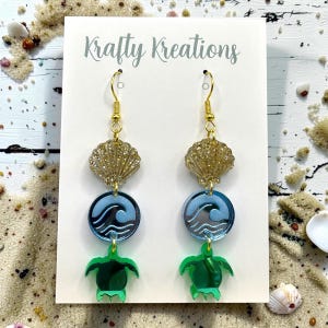 May include: Dangle earrings on a white Krafty Kreations display card. Each earring features a gold glitter seashell, a light blue circular charm with a wave design, and a green sea turtle charm, all connected by gold-toned metal hooks.