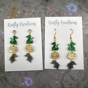 May include: Two pairs of dangle earrings. Each earring features a green glitter dragon, a stack of gold books, and a gray creature. The earrings are on white cards with the text "Krafty Kreations".