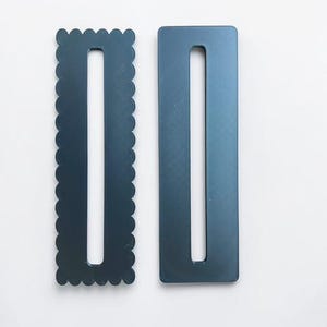May include: Two dark gray rectangular belt buckles. One buckle features a scalloped edge, while the other has a smooth, straight edge. Both buckles contain a long, narrow vertical slot in the center for threading a belt strap through.