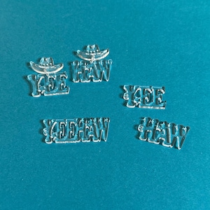以下が含まれることがあります： 「YEE HAW」の文字とカウボーイハットのデザインが施された透明アクリルカットアウト。カットアウトは様々なサイズと配置で、クラフトや装飾に最適です。背景は無地のティール色です。